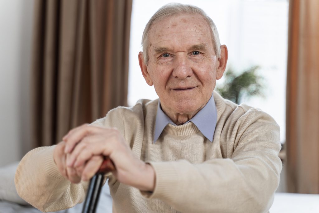 Elderly man in a warm sweater with a walking cane indoors at BeeHive Homes of Rowlett, featuring soft lighting and a plant.