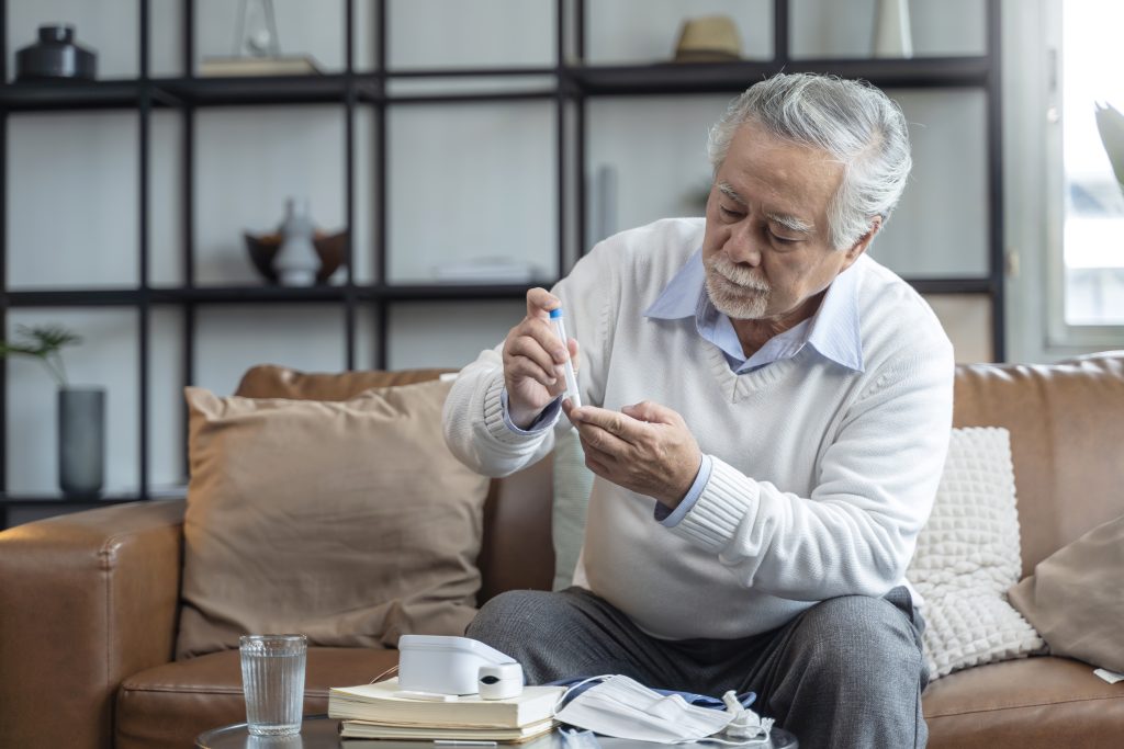 Elderly man on a couch holding a thermometer, with a glass of water and medical device at BeeHive Homes of Rowlett.