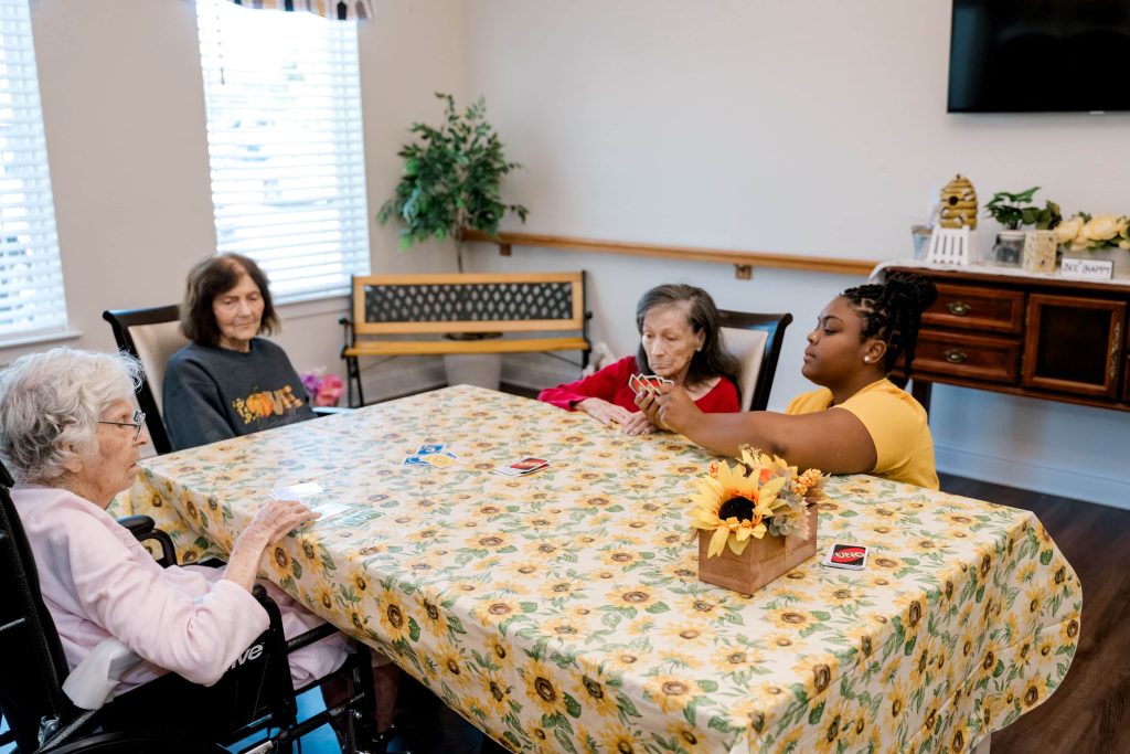 Elderly women and a young woman play cards at a bright table in a senior care facility setting.