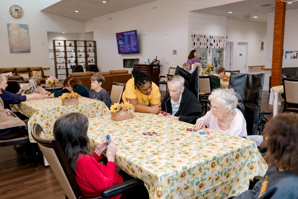 Elderly residents engaged in activities at BeeHive Homes, featuring tables, playing cards, and a bright interior.