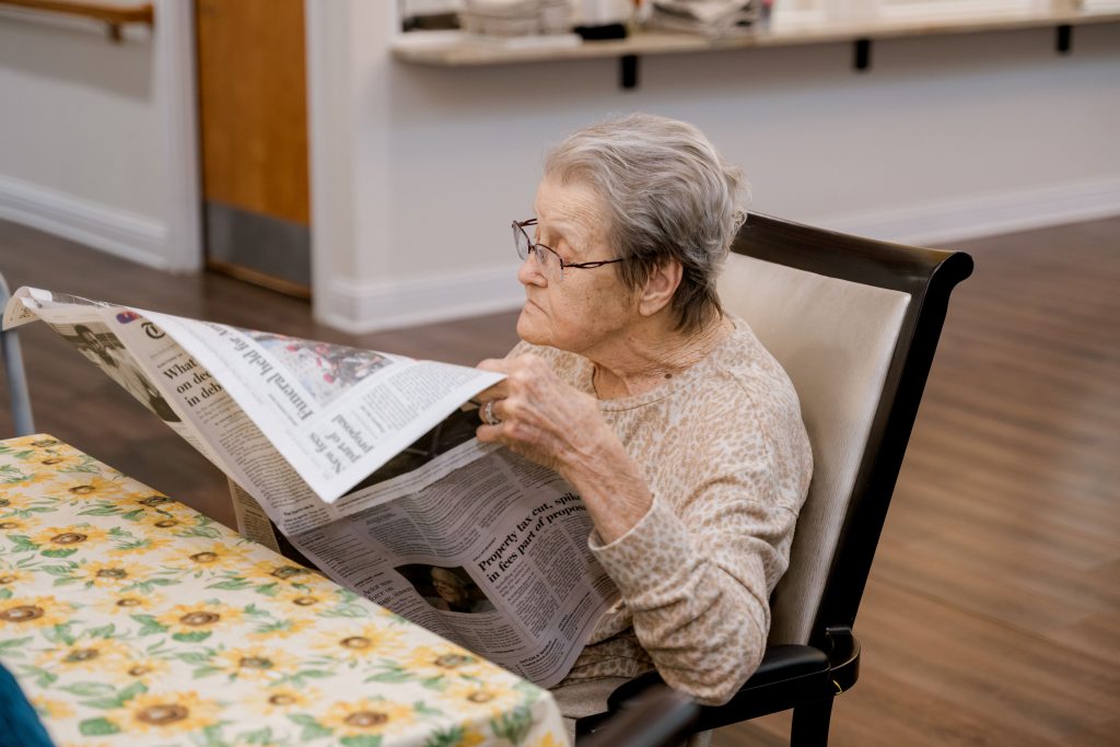 Elderly woman reading a newspaper at a table with a floral tablecloth, highlighting senior living and memory care themes.