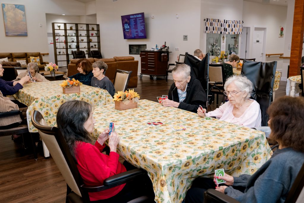 Elderly residents at BeeHive Homes playing Uno at tables with floral tablecloths in a bright, communal setting.